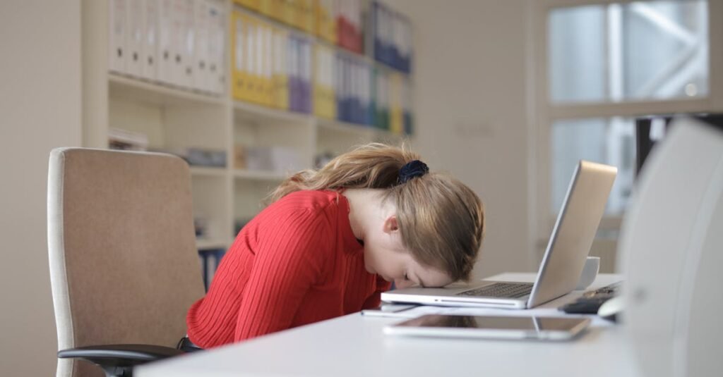 A tired woman in a red sweater leans her head on a desk with a laptop, symbolizing workplace fatigue.