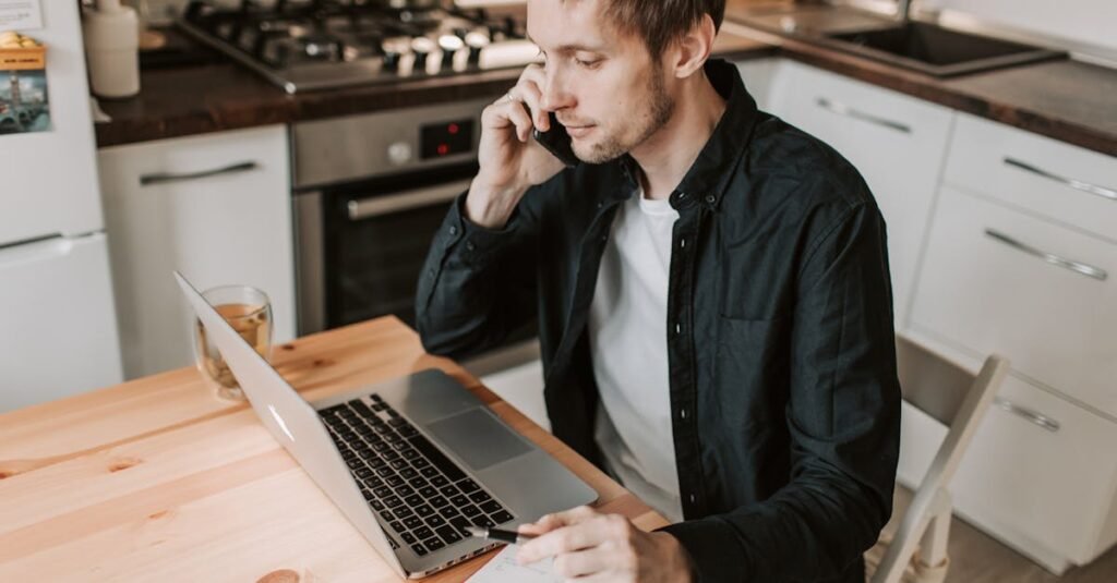 A young man works from home on his laptop, communicating over the phone in a serene kitchen setting.