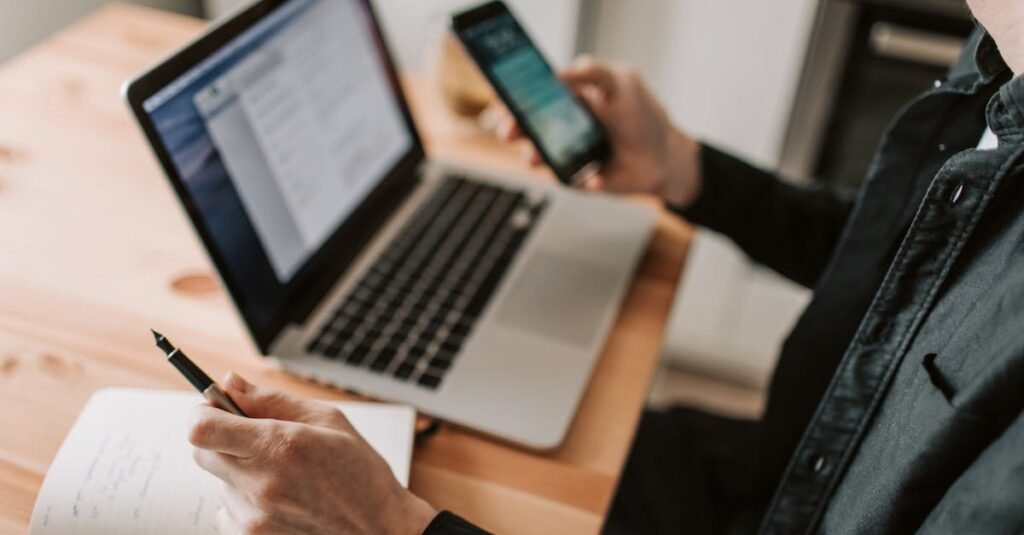A person multitasking with a laptop and smartphone at a wooden desk.